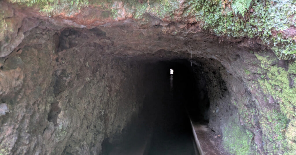 Looking through a long, low tunnel, with a levada flowing through it and a narrow path to the side.