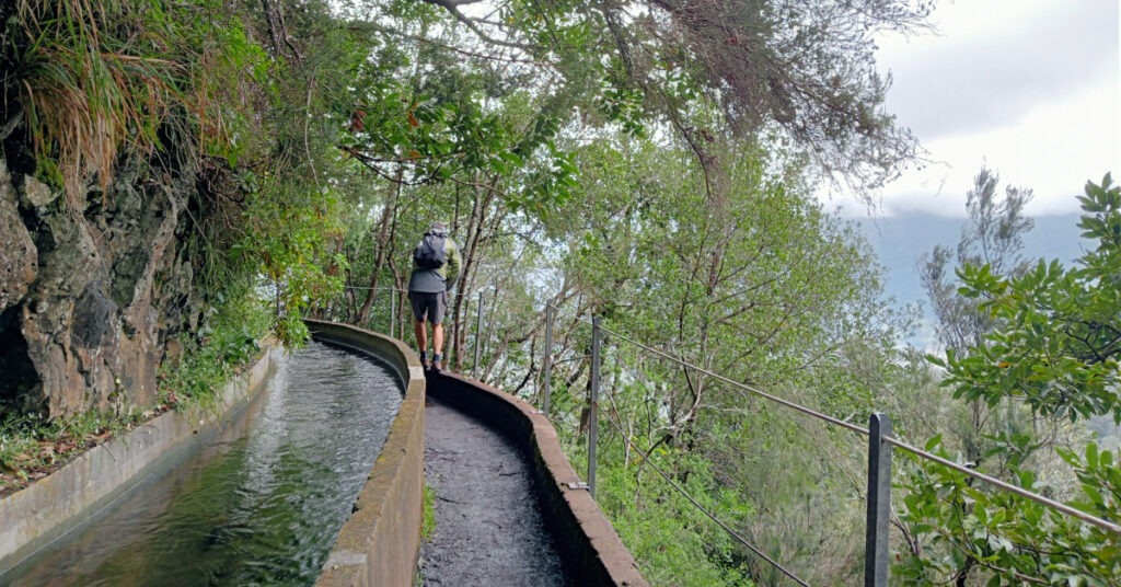 John walks along the narrow stone edge of a path. There is the levada and the flooded path on his left, a sheer drop on his right.